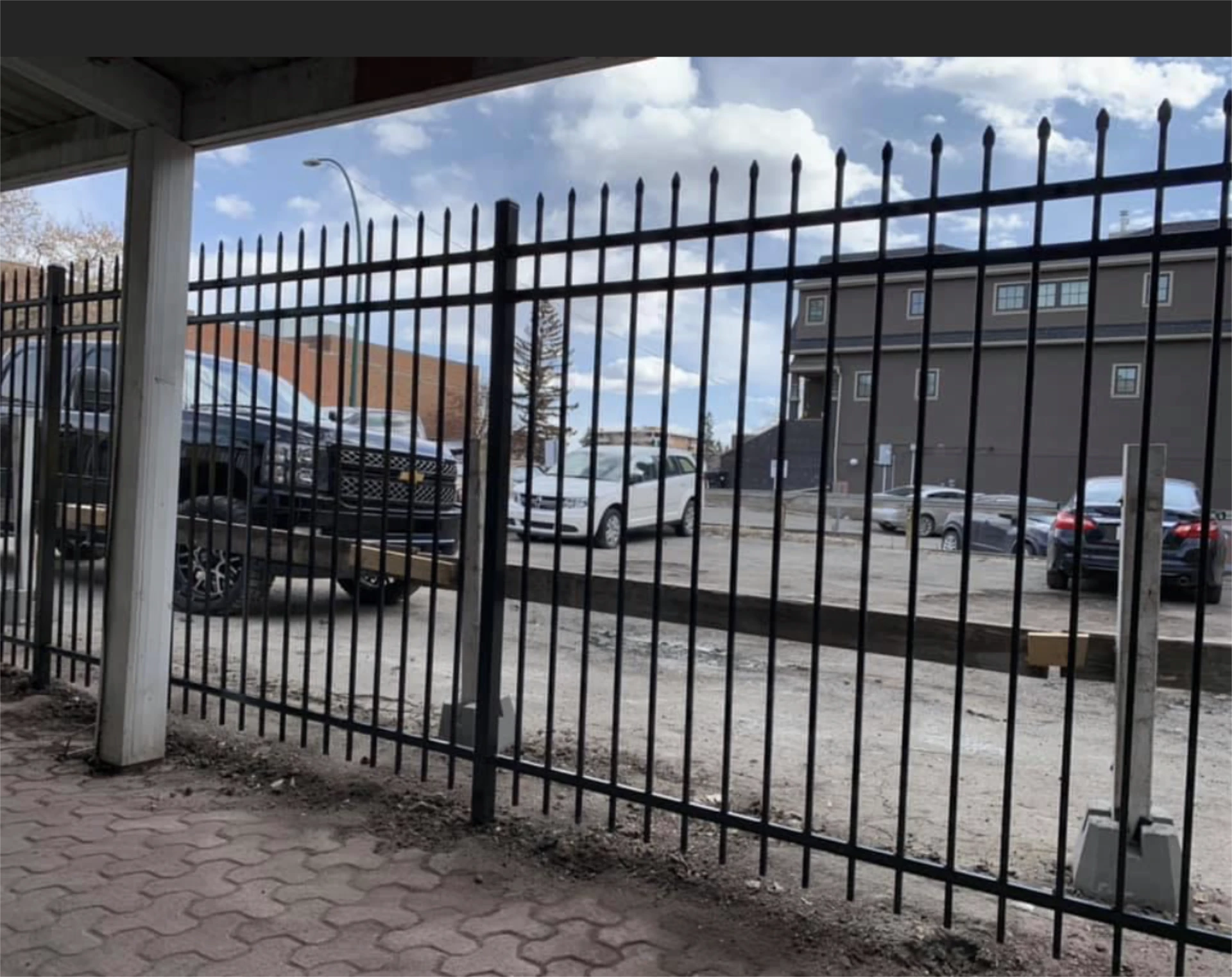 Black metal fence enclosing a gravel parking lot with parked cars and a building in the background.