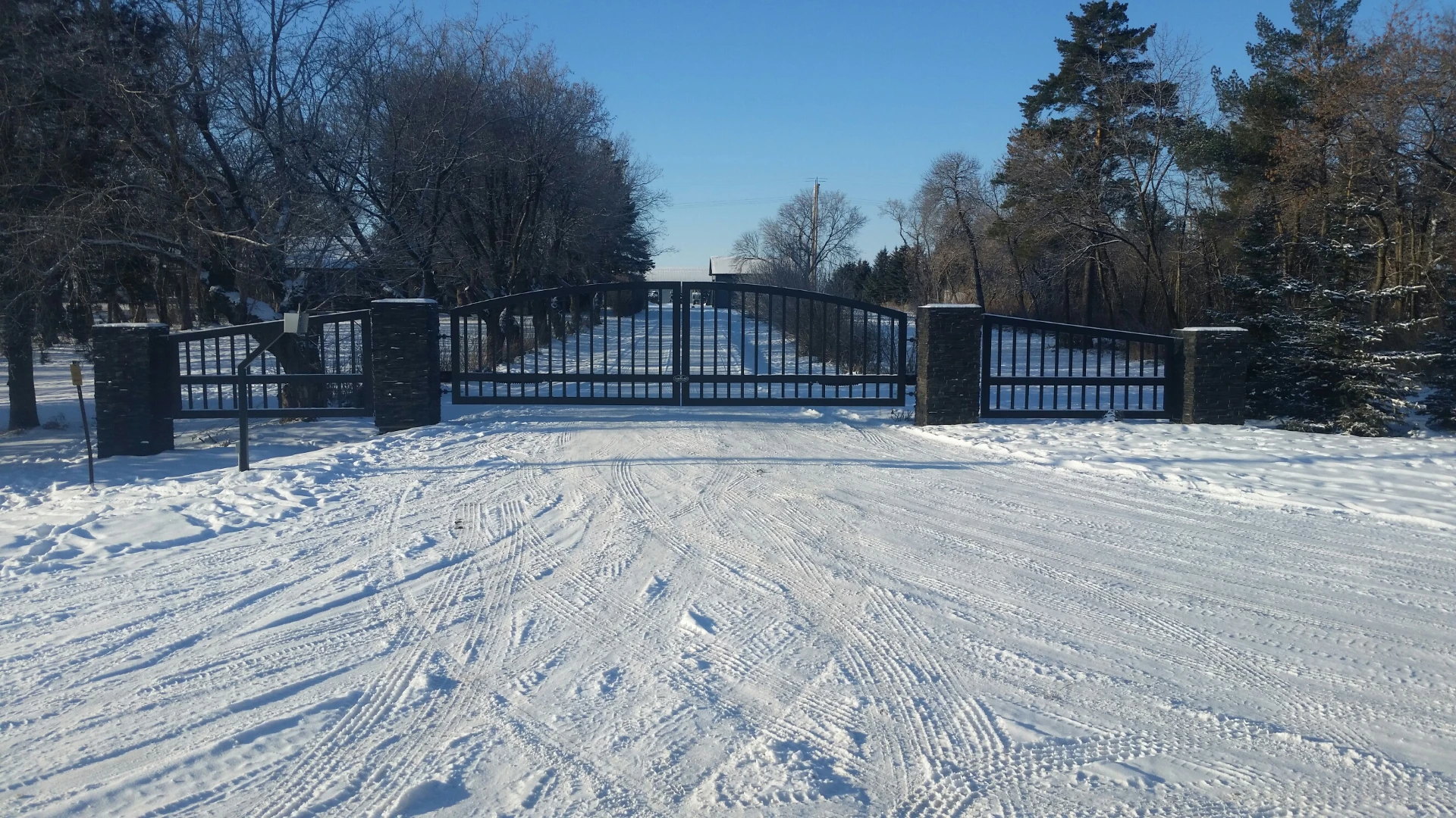 Snow covered driveway leading to a closed metal gate surrounded by trees under a clear blue sky.